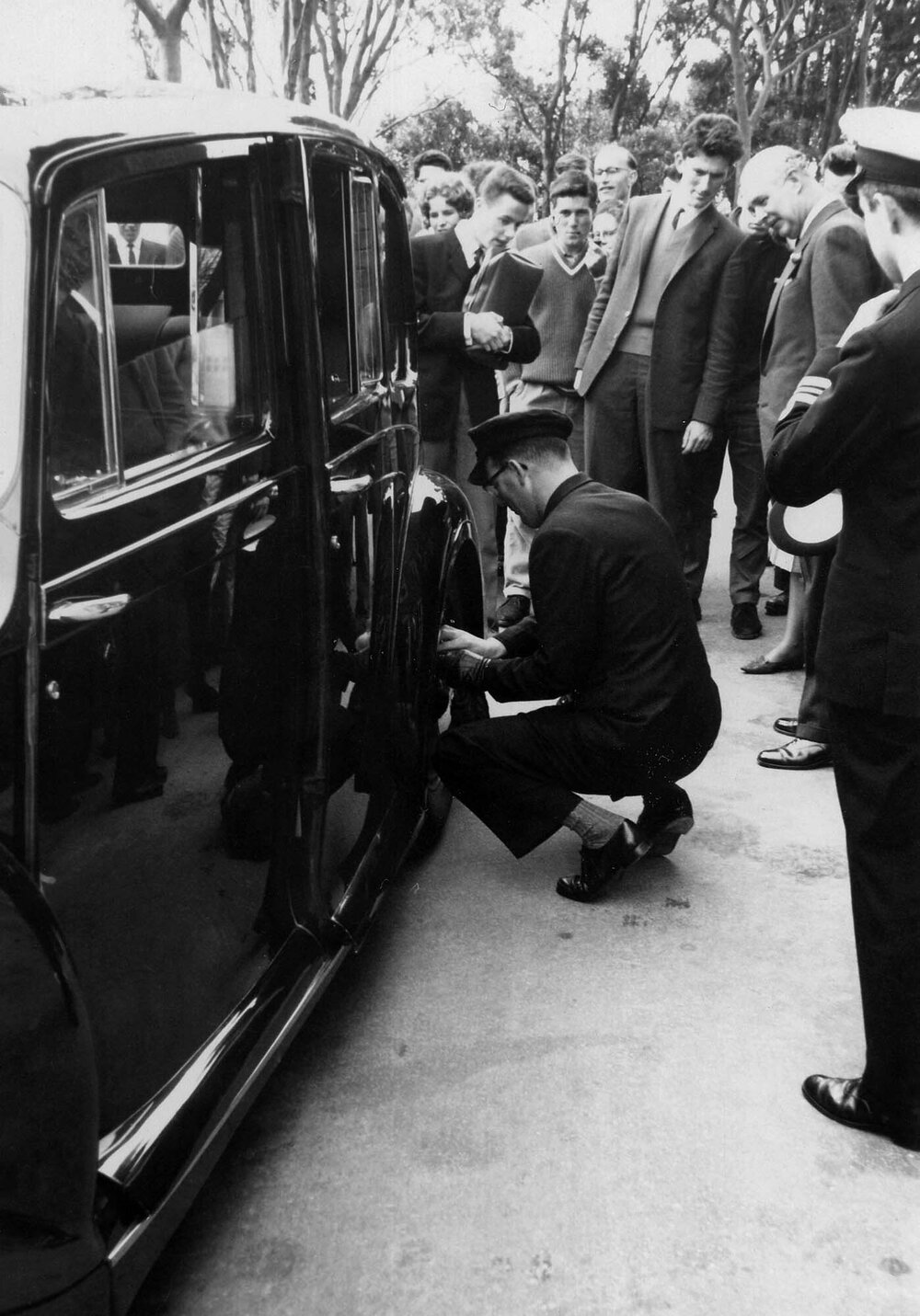 Governor General's chauffer removing coins from car hubcaps